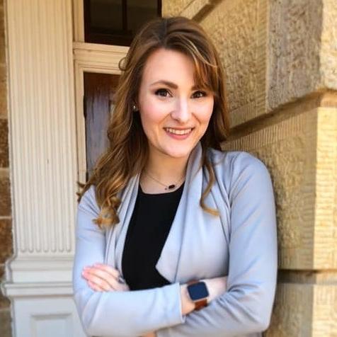 Amy stands smiling with her arms crossed. She is standing in front of a sandstone-like brick wall.